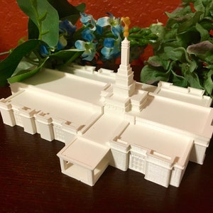 Może przedstawiać: Biały model 3D świątyni wydrukowany w technologii 3D z wysoką wieżą i skomplikowanymi detalami. Model stoi na drewnianej powierzchni z zielonymi roślinami w tle.