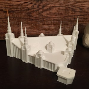 Può includere: Un modello architettonico bianco di un edificio con più guglie e una base rettangolare. Il modello presenta dettagli, tra cui più torri e una piccola guglia color oro. Il modello è ambientato su uno sfondo di legno scuro.