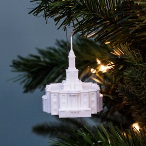 May include: A white architectural ornament, resembling a temple, hangs from a Christmas tree. The ornament has a detailed facade with columns and a spire. The background is a blurred blue-green with Christmas tree branches and lights.