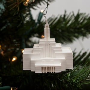May include: White 3D printed ornament shaped like a church building, hanging from a Christmas tree branch.