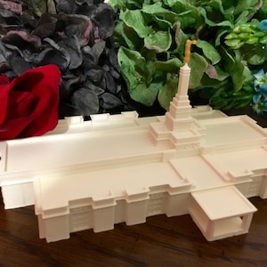 May include: A white miniature replica of a temple with a gold spire. The replica is sitting on a wooden surface with flowers in the background.