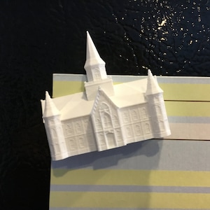 May include: White 3D printed model of a church building with a tall spire and multiple windows. The model is on a striped background.