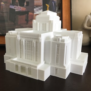 May include: A white 3D printed model of a building with a gold spire on top. The building has many windows and a detailed facade.