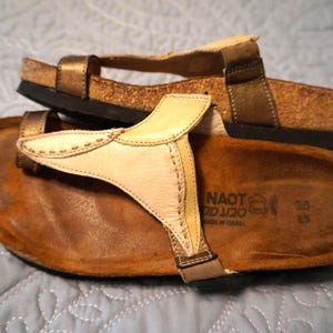 May include: A pair of Naot sandals featuring a thong-style design with a cream-colored leather upper and bronze-toned straps. The footbed is made of cork and brown suede, with the brand name and size 38 printed clearly on the heel area of the sole.