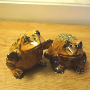May include: A pair of ceramic toad salt and pepper shakers featuring a glossy brown and orange glaze with textured green spots on the back. Each toad has a wide mouth, dark brown limbs, and small holes on the top of the head for dispensing seasoning.