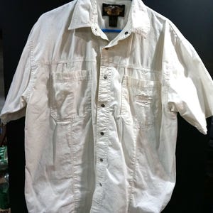 Może przedstawiać: Biała koszula z krótkim rękawem zapinana na guziki, z dwiema kieszeniami na piersi i metką z logo "Harley Davidson" z przodu.