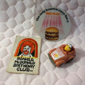 Op de afbeelding: Een vintage McDonald's speelgoedauto met een gele Ronald McDonald-figuur achter het stuur. De auto is bruin met een geel M-logo en de tekst "Early Bird" aan de zijkant. Er is ook een rood, wit en geel geborduurd pleister met Ronald McDonald's gezicht en de tekst "Ronald McDonald Birthday Club". Een witte sticker met de tekst "The only known cure for a Big Mac Attack" en een afbeelding van een Big Mac is ook inbegrepen.