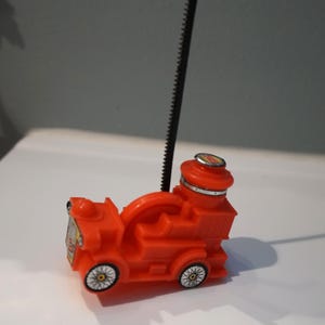 May include: A bright orange toy train with black wheels and a tall, black, ridged antenna. The train has a detailed design, including a smokestack and a front grill. The toy is set against a white surface.