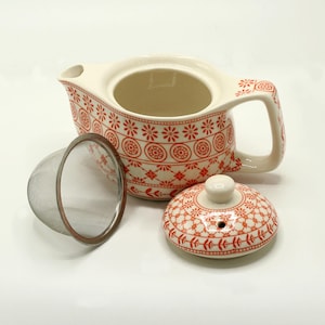 May include: A cream ceramic teapot with a red floral design, lid, and a stainless steel tea infuser. The teapot has a handle and a spout. The lid has a small knob. The tea infuser is a mesh basket.