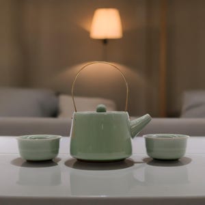 May include: A light green ceramic teapot with a gold handle and lid, along with two matching teacups. The teapot and cups are sitting on a white table.