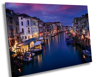 May include: A view of a canal in Venice, Italy at dusk. The water is calm and reflects the colorful buildings on either side. There are many boats docked along the canal, and the sky is a beautiful mix of pink and purple.