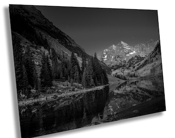May include: Black and white landscape photograph of a mountain range reflected in a calm lake. The image features tall, snow-capped peaks, lush evergreen forests, and a dark, starry sky. The composition highlights the natural beauty of the wilderness.