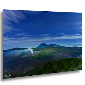 Può includere: Una fotografia paesaggistica di una regione vulcanica di notte sotto un cielo blu stellato. Un cratere fumante si trova in primo piano accanto a una montagna verde a forma di cono. In lontananza, una vetta più grande si staglia all'orizzonte.