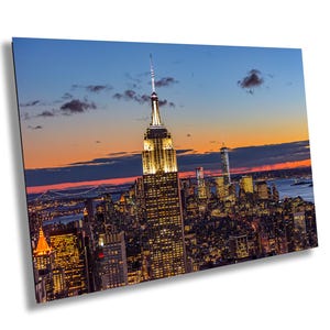 Könnte beinhalten: Eine Luftaufnahme der Skyline von New York City in der Abenddämmerung, mit dem Empire State Building im Zentrum des Bildes beleuchtet. Der Himmel ist leuchtend orange und rosa, mit Wolken in der Ferne. Die Lichter der Stadt funkeln darunter.