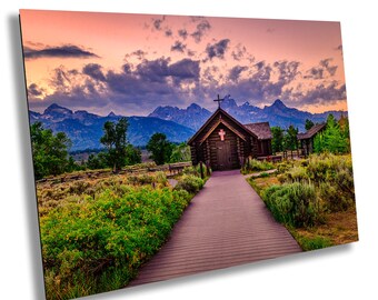 May include: A wooden chapel with a cross on the roof stands in a field with a wooden walkway leading to it. The chapel is surrounded by green grass and trees. The background features a mountain range with a pink and purple sunset sky.