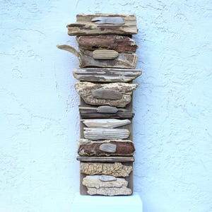May include: A vertical wall art piece constructed from stacked pieces of weathered driftwood in various shades of brown, grey, and tan. The driftwood is arranged in a layered, textured column with smaller, smooth, rounded stones attached to the centre.