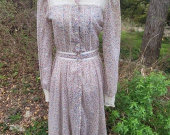 Puede incluir: Vestido vintage con estampado floral, mangas largas y botones delanteros. El vestido presenta un cuello y puños de encaje color crema, con un ribete de encaje a juego en el dobladillo. El vestido se exhibe en un maniquí al aire libre.