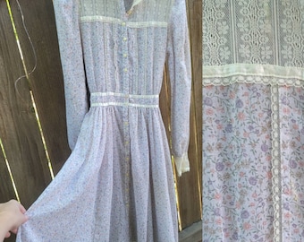 Puede incluir: Vestido vintage con estampado floral en tonos lavanda, rosa y verde. El vestido presenta mangas largas, una botonadura frontal y delicados detalles de encaje en el cuello, el corpiño y el dobladillo. Confeccionado en tejido ligero y semitransparente.
