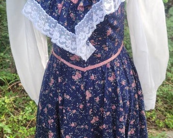Puede incluir: Vestido vintage azul marino con estampado floral, ribete de encaje blanco y cuello alto. El vestido presenta un panel de terciopelo con botones y detalles en rosa. Las mangas largas son blancas.