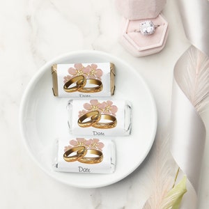 May include: Three white chocolate bar wrappers with pink flowers and gold rings. The wrappers have the word "Date" printed on them.