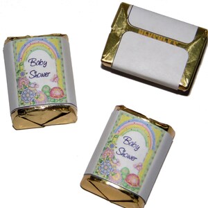 May include: Three small chocolate bars wrapped in gold foil with a white paper label. The label has a rainbow, flowers, and the text "Baby Shower".