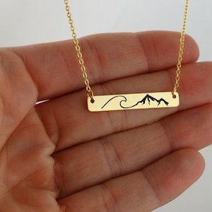 May include: Gold chain necklace with a horizontal bar pendant featuring a black silhouette of a wave and mountain range.