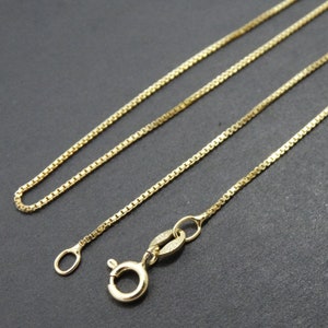 May include: A gold-colored box chain necklace with a lobster clasp and a small loop at the end. The chain is made of small, square links and is displayed against a dark background. The necklace is a piece of jewelry.