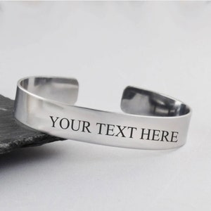 May include: A silver cuff bracelet with the engraved text "YOUR TEXT HERE". The bracelet has a simple, open design and is resting on a dark, textured surface. The metal has a polished, reflective finish.