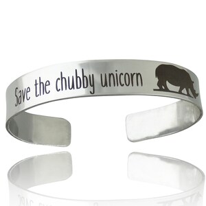 May include: A silver cuff bracelet with the phrase "Save the chubby unicorn" engraved on the front. A silhouette of a rhino is on the right side of the bracelet. The bracelet is open-ended and has a reflective surface.