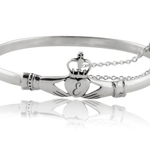 May include: A silver-toned Claddagh bangle bracelet featuring two hands holding a heart topped with a crown. The heart is engraved with the letter E. The bracelet includes a hinged opening and a delicate safety chain attached to the side.