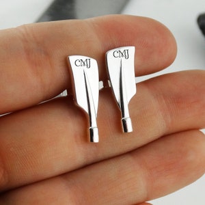 May include: A pair of silver cufflinks shaped like oars. Each cufflink has the initials "CMJ" engraved on the blade. The cufflinks are displayed against a neutral background.