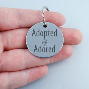 May include: Silver round dog tag with the text "Adopted and Adored" engraved on it.