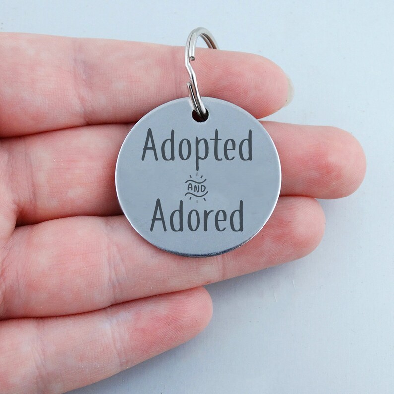 May include: Silver round dog tag with the text "Adopted and Adored" engraved on it.