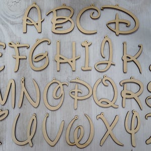 May include: A complete alphabet of wooden letters, each with a small hole at the top for hanging. The letters are in a natural wood colour, with a variety of fonts, including a stylized Disney font. The letters are arranged in rows and columns.