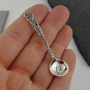 May include: A small silver-toned spoon featuring an intricate floral relief pattern along the handle. The bowl of the spoon is smooth and features a dark, cursive letter G engraved in the center, providing a clear contrast against the metallic surface.