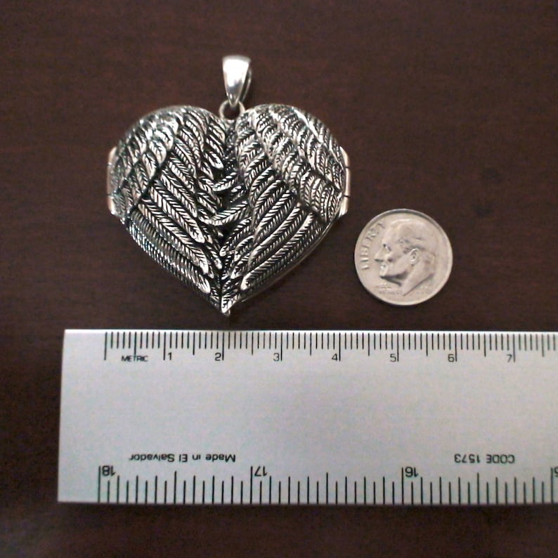 May include: Silver heart-shaped locket featuring a detailed design of angel wings. The locket measures approximately 5 cm in diameter.