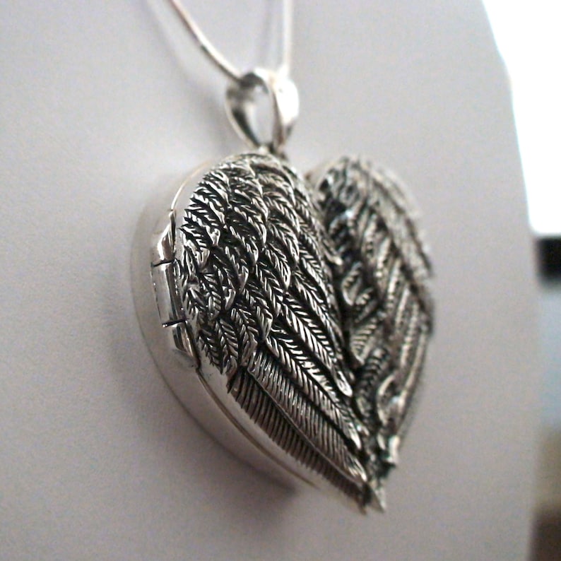 May include: Silver heart-shaped locket with angel wings. The locket is open and the inside is visible.