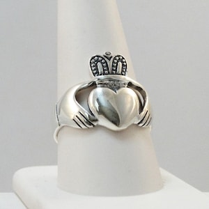 May include: A polished silver Claddagh ring featuring a central heart held by two hands, topped with a detailed crown. The metal surface is smooth and reflective, showcasing the traditional design elements of the heart, hands, and crown clearly.