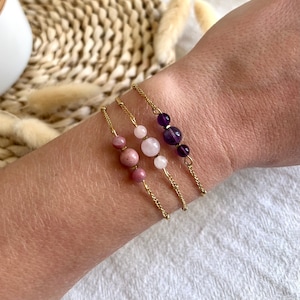 Peut inclure: Trois bracelets délicats en chaîne dorée, chacun orné d'une combinaison différente de pierres précieuses. L'un présente des pierres roses et roses, un autre des pierres roses claires et blanches, et le troisième des pierres violettes. Les bracelets sont portés sur un poignet.