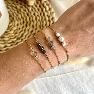 May include: Four gold chain bracelets, each featuring different gemstone beads. The bracelets are displayed on a wrist, showcasing the variety of colours and textures of the beads. The beads include clear, grey, and white stones.