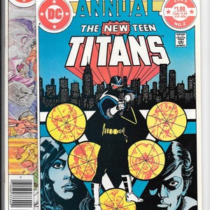 May include: Comic book cover for "Annual The New Teen Titans" featuring a costumed character aiming a weapon. The cover is predominantly blue, red, and yellow, with a cityscape backdrop. The comic is from 1983 and has a price tag of $1.00.