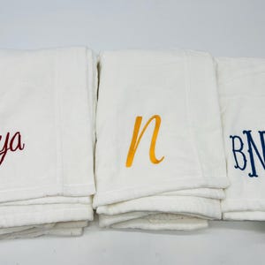 May include: Three white blankets with embroidered lettering. One blanket has the name "Mya" in red script, another has the letter "n" in yellow, and the third has the initials "BNE" in blue.