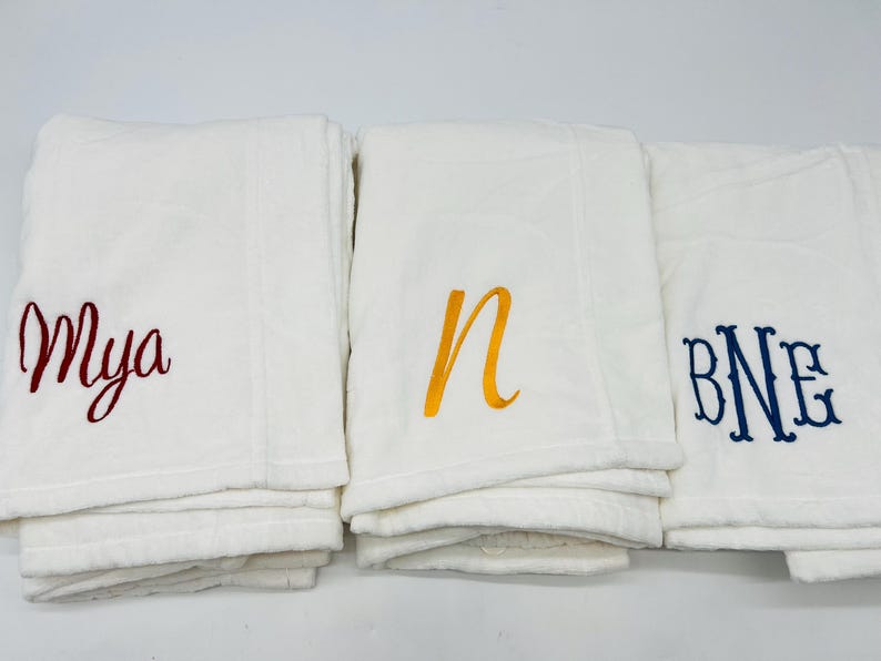 May include: Three white blankets with embroidered lettering. One blanket has the name "Mya" in red script, another has the letter "n" in yellow, and the third has the initials "BNE" in blue.