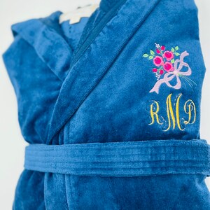 Puede incluir: Una bata de terciopelo azul con un dise&ntilde;o floral bordado en blanco y el monograma "RMG" en hilo dorado.