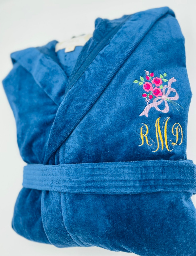 Puede incluir: Una bata de terciopelo azul con un dise&ntilde;o floral bordado en blanco y el monograma "RMG" en hilo dorado.
