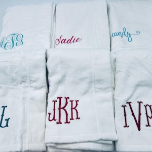 May include: White bath towels with embroidered names and monograms in various colors. The towels feature names like "Sadie" and "candy", and monograms such as "GBL" and "IVA". The embroidery colors include teal, pink, and burgundy.