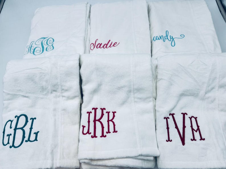 May include: White bath towels with embroidered names and monograms in various colors. The towels feature names like "Sadie" and "candy", and monograms such as "GBL" and "IVA". The embroidery colors include teal, pink, and burgundy.