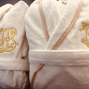May include: Two white bathrobes with embroidered initials. The robe on the left has the initials "JB" embroidered in gold thread. The robe on the right has the initials "TB" embroidered in gold thread.