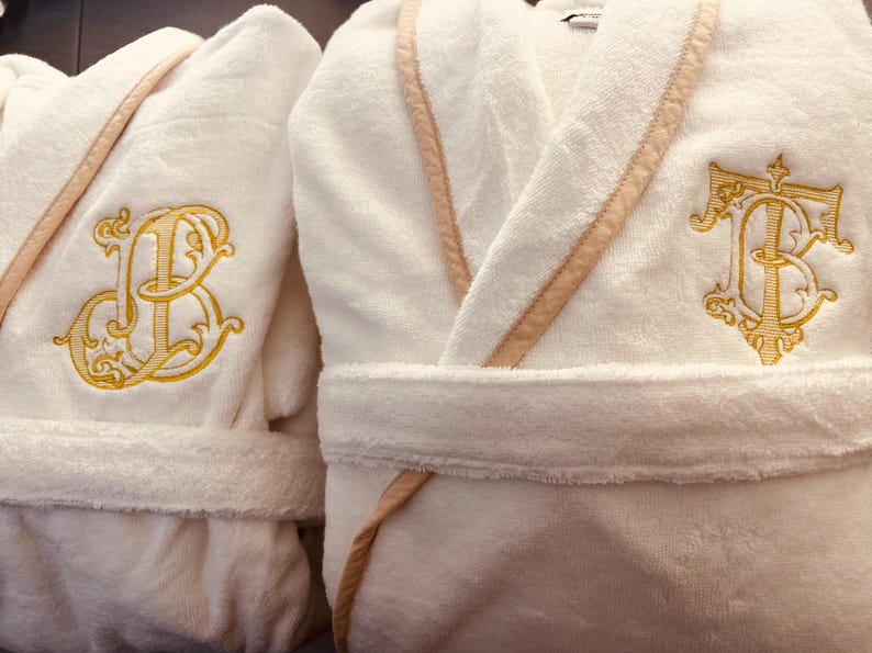 May include: Two white bathrobes with embroidered initials. The robe on the left has the initials "JB" embroidered in gold thread. The robe on the right has the initials "TB" embroidered in gold thread.