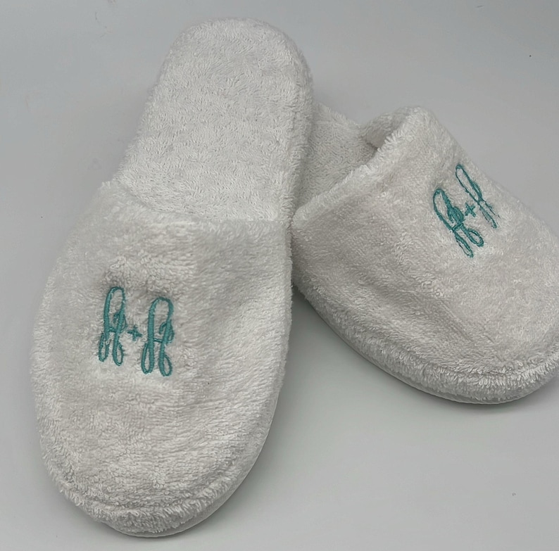 May include: A pair of white terry cloth slippers. Each slipper has a teal embroidered monogram. The slippers are soft and plush, perfect for lounging. The monogram appears to be the letters "A" and "R" intertwined.